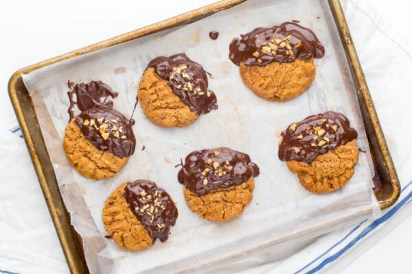 Six Chocolate-dipped peanut butter cookies are sitting on a baking sheet that has been lined with parchment paper.
