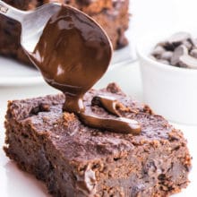 A spoon hovers over these chocolate banana brownies, drizzling melty chocolate over the top. A bowl of chocolate chips sits on the right and more slices of brownies are in the background.