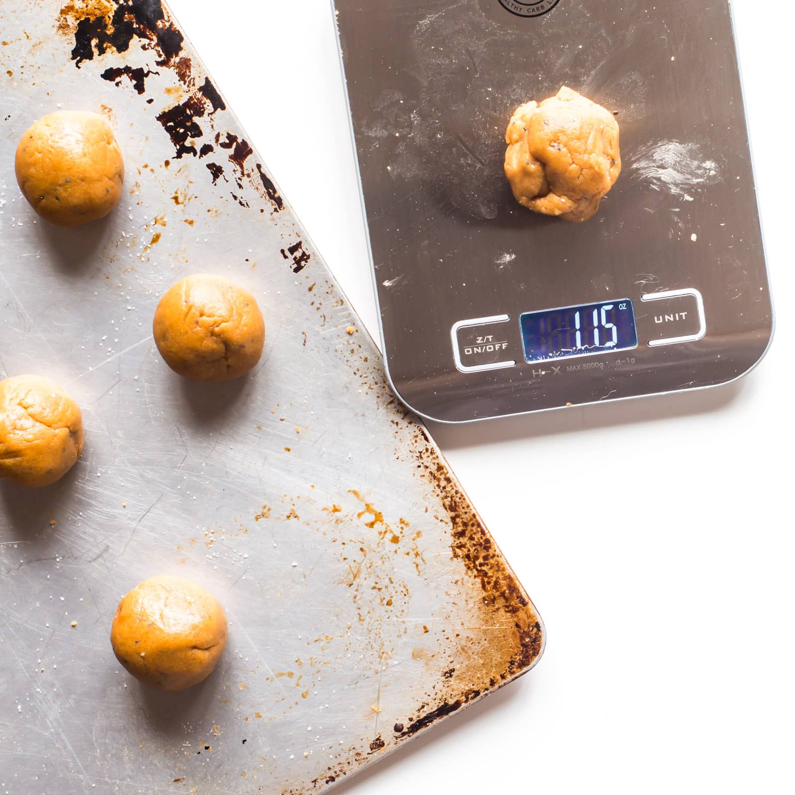 The cookie dough for these vegan peanut butter balls sit on a baking pan. A pinch of the dough is sitting on a kitchen measuring tool to help weight cookies to make them equally sized.