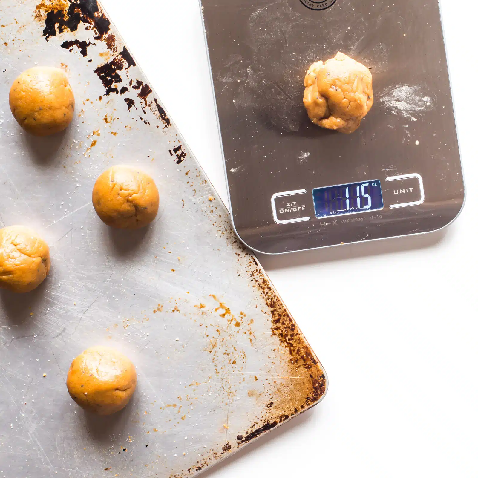 The cookie dough for these vegan peanut butter balls sit on a baking pan. A pinch of the dough is sitting on a kitchen measuring tool to help weight cookies to make them equally sized.
