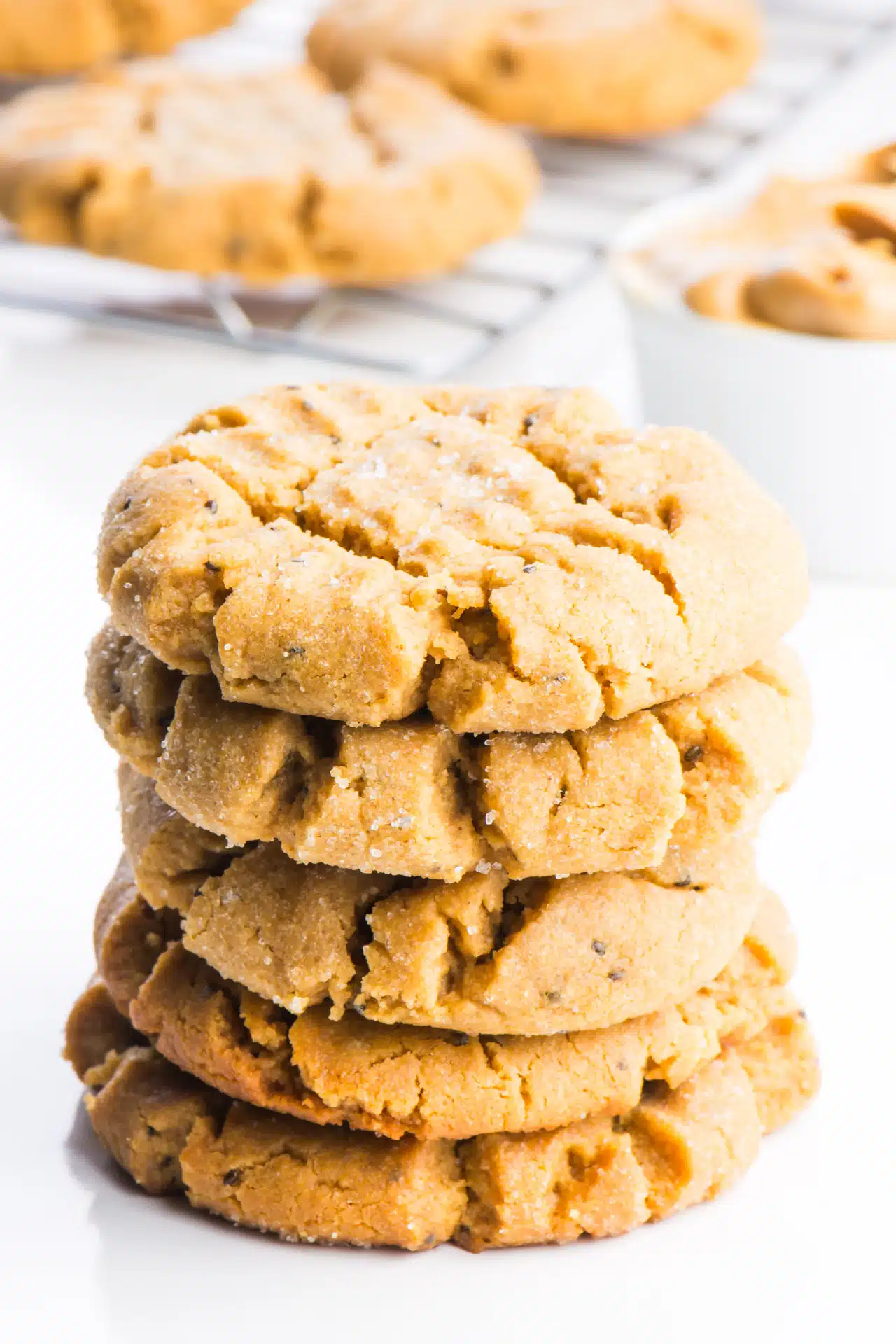 A stack of vegan peanut butter cookies sits in the foreground. Behind them is a white container full of peanut butter and a wire rack with more cookies cooling after being pulled from the oven.