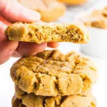 A square-cropped image of a stack of vegan peanut butter cookies and a hand holding one with a bite taken out.