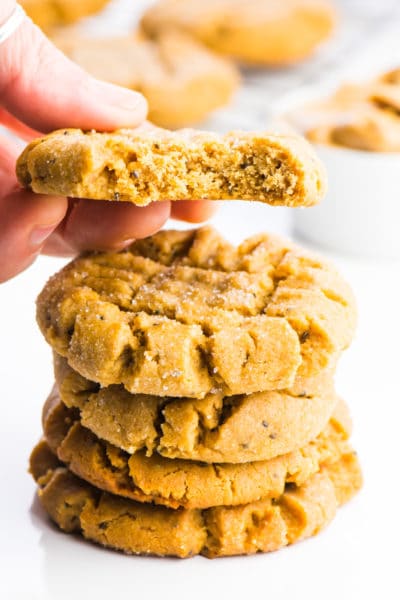 A stack of vegan peanut butter cookies is sitting in the foreground. A hand is holding a cookie right above the stack and there's a bite taken out of that cookie.
