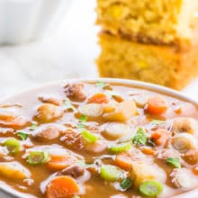 A bowl of pinto bean soup is front in center in this photo. The light is shining on all the ingredients in the bowl, including the slices of carrots, potatoes, and beans. There is a stack of cornbread behind the soup, and a bowl of dried pinto beans behind that.