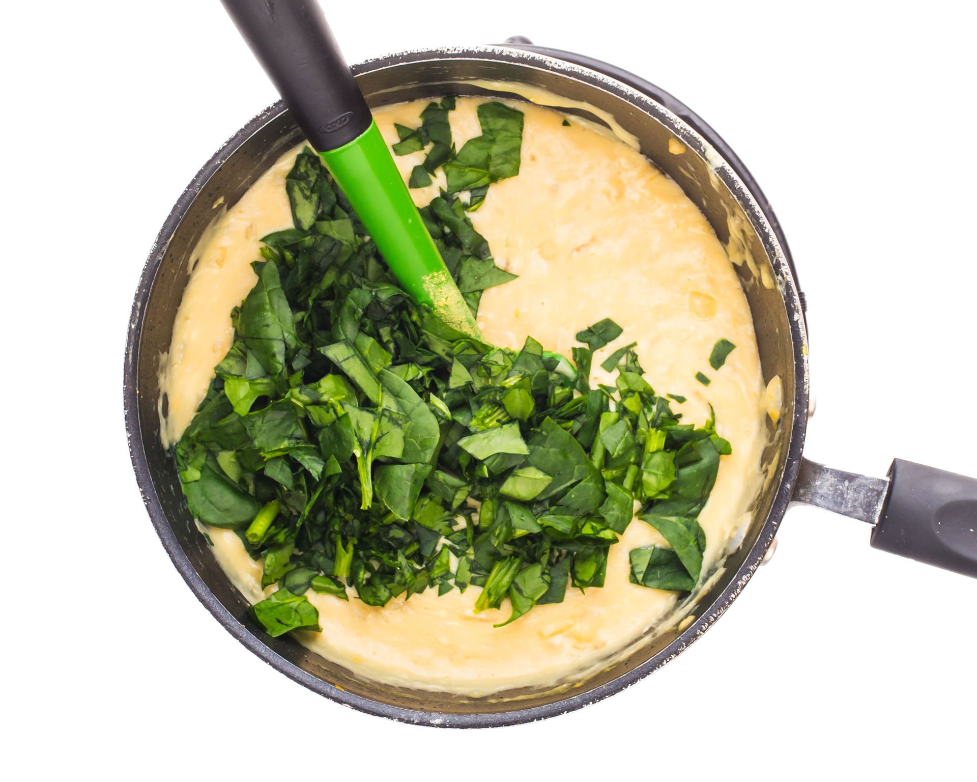 A saucepan with melted vegan cheese and chopped spinach and a green spatula.