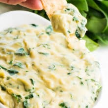 A hand has scooped out some vegan spinach artichoke dip on a chip. The full bowl of dip is below it, and fresh spinach is behind it.