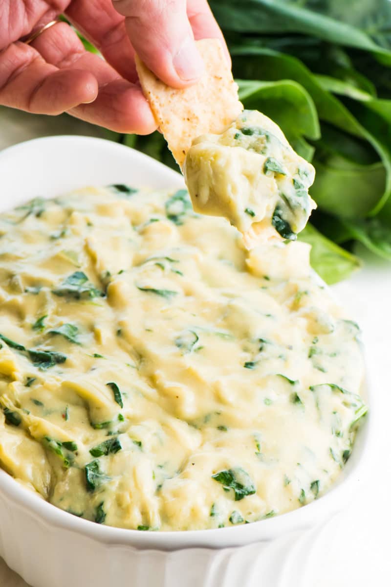 A hand has scooped out some vegan spinach artichoke dip on a chip. The full bowl of dip is below it, and fresh spinach is behind it.