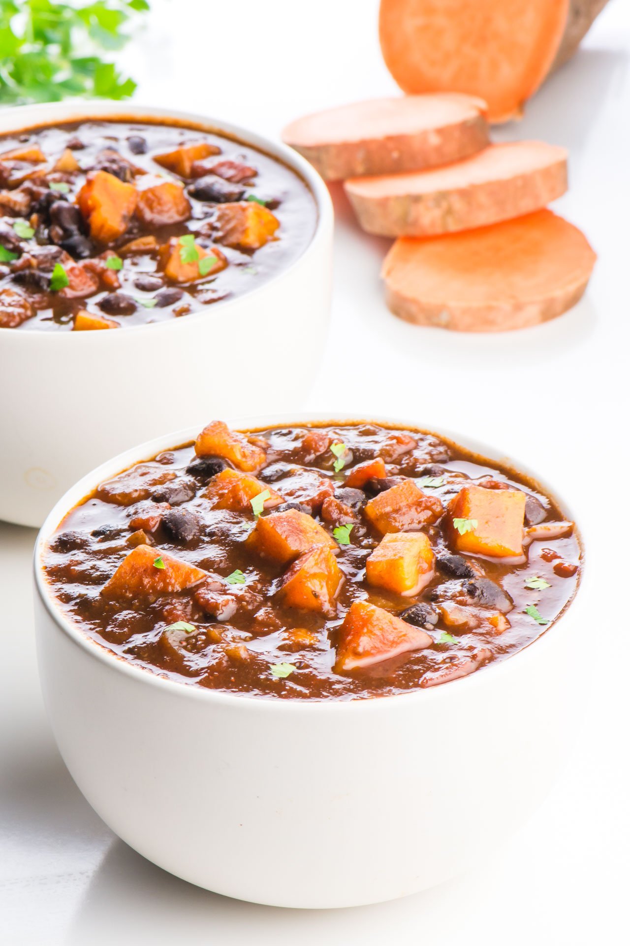 Two bowls of sweet potato chili are in this photo. There is one bowl in the front and another behind it. There are slices of sweet potato and a sprig of parsley behind them.