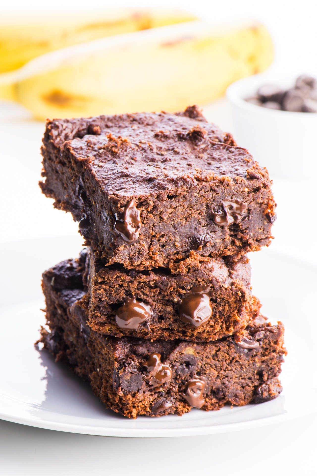 Three chocolate banana brownies sit stacked on each other. There are bits of melted chocolate chips bursting from the sides of each brownies. A white bowl full of dark chocolate chips sits behind the brownies and some yellow bananas are behind that.