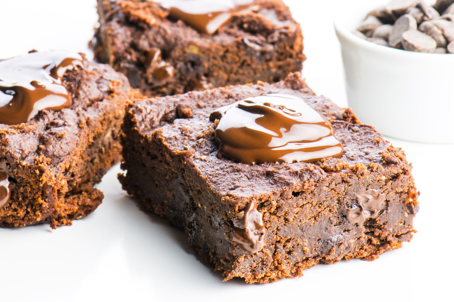 Three slices of chocolate banana brownies sit next to each other, each with a dollop of melted chocolate over the top. A white bowl full of chocolate chips sits nearby.