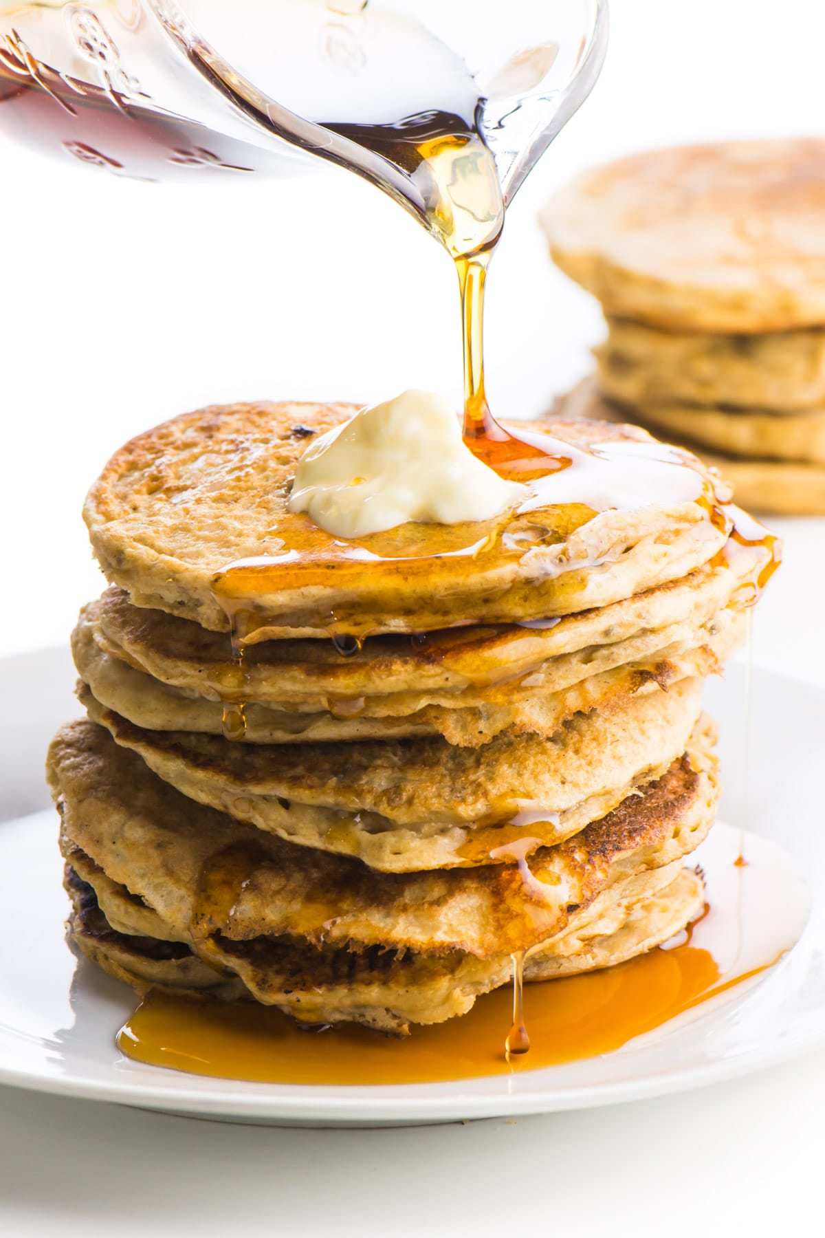 A stack of these pancakes without eggs is sitting on a plate. The top one has a pat of melty vegan butter. A glass pitcher is pouring maple syrup and it is dropping down the sides of the pancakes. There are more pancakes in the background.