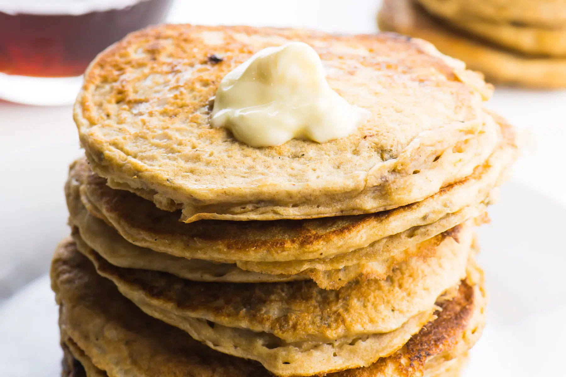 A stack of pancakes sit on a plate with a pat of melted vegan butter on the top. There are more pancakes in the background and a glass pitcher with maple syrup waits to be poured over the top.
