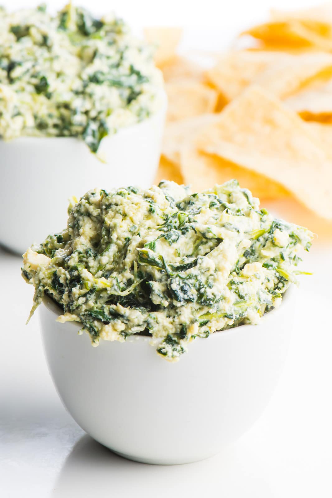 Two white dishes hold vegan spinach dip, one in front of the other. A pile of tortilla chips sit in the background.