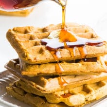 A glass pouring cup is pouring maple syrup over a stack of whole wheat vegan waffles.