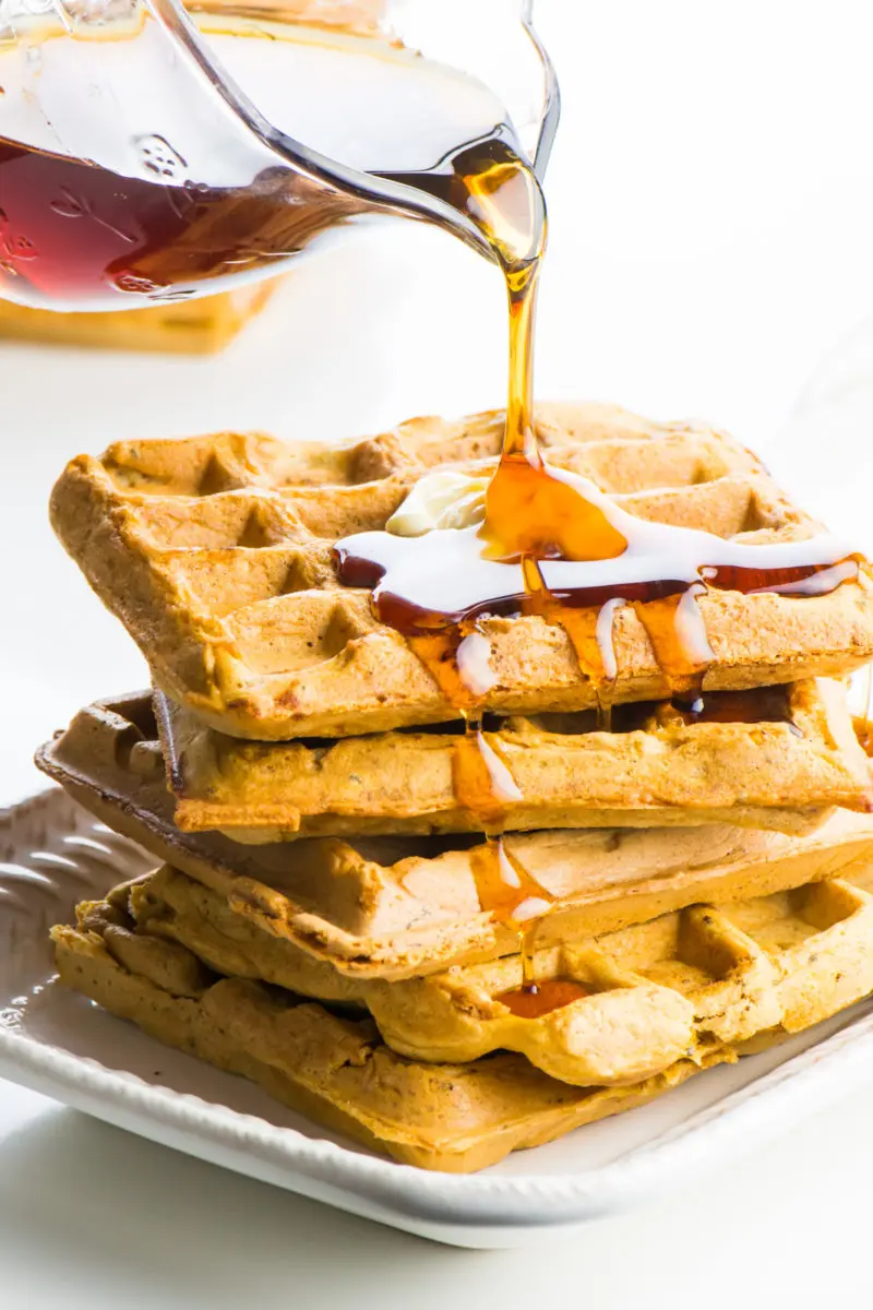 A glass pouring cup is pouring maple syrup over a stack of whole wheat vegan waffles.