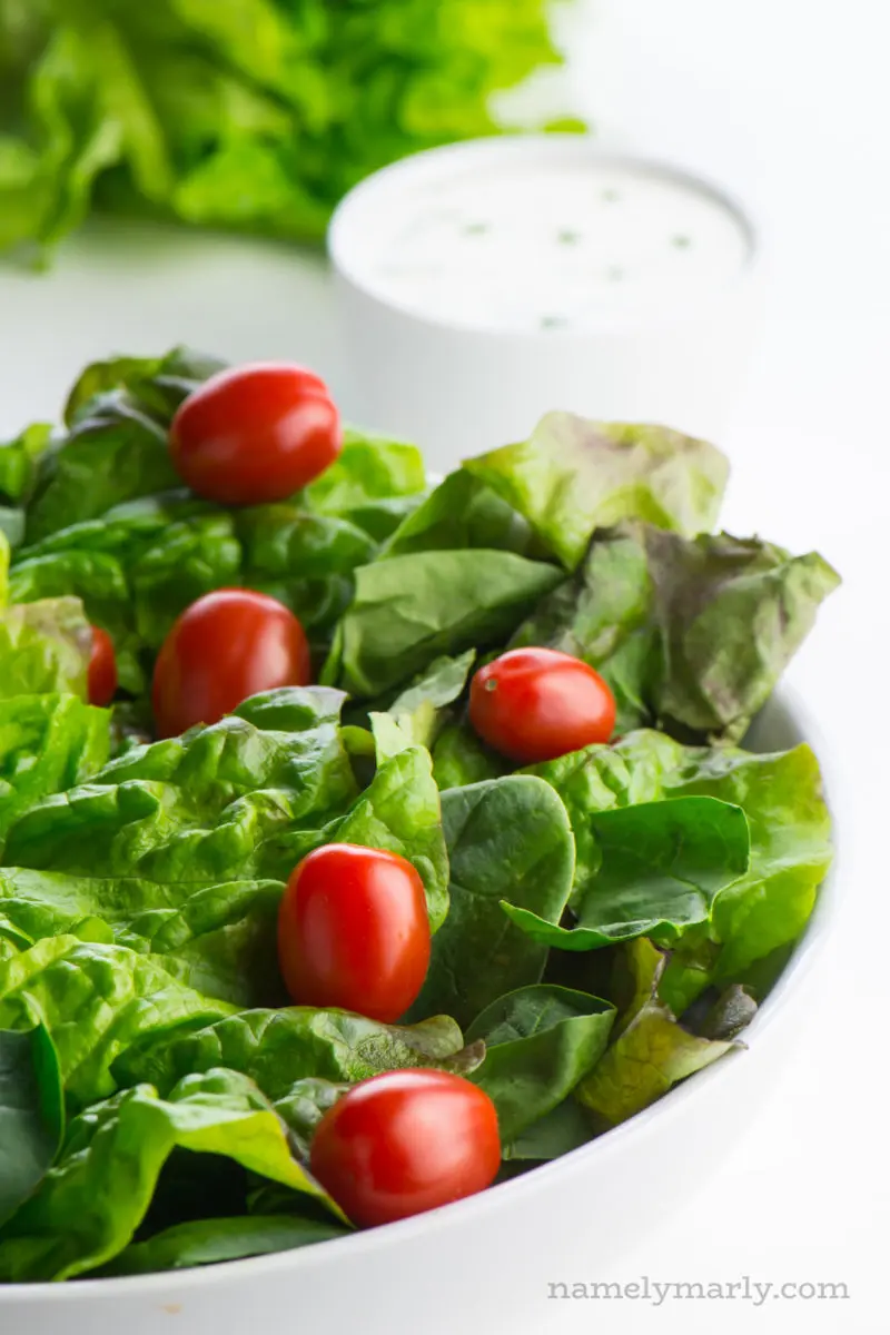 A white bowl full of bright, leafy greens sits in the foreground. A bowl of dressing is behind it.