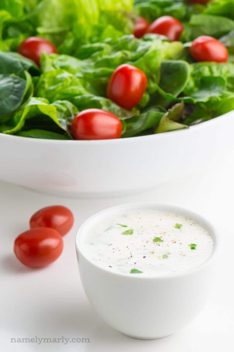 A bowl of vegan ranch dressing sits in front of a salad bowl full of greens and cherry tomatoes.