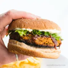 A hand holds a lentil burger, showing off greens and mayo on a whole wheat bun.