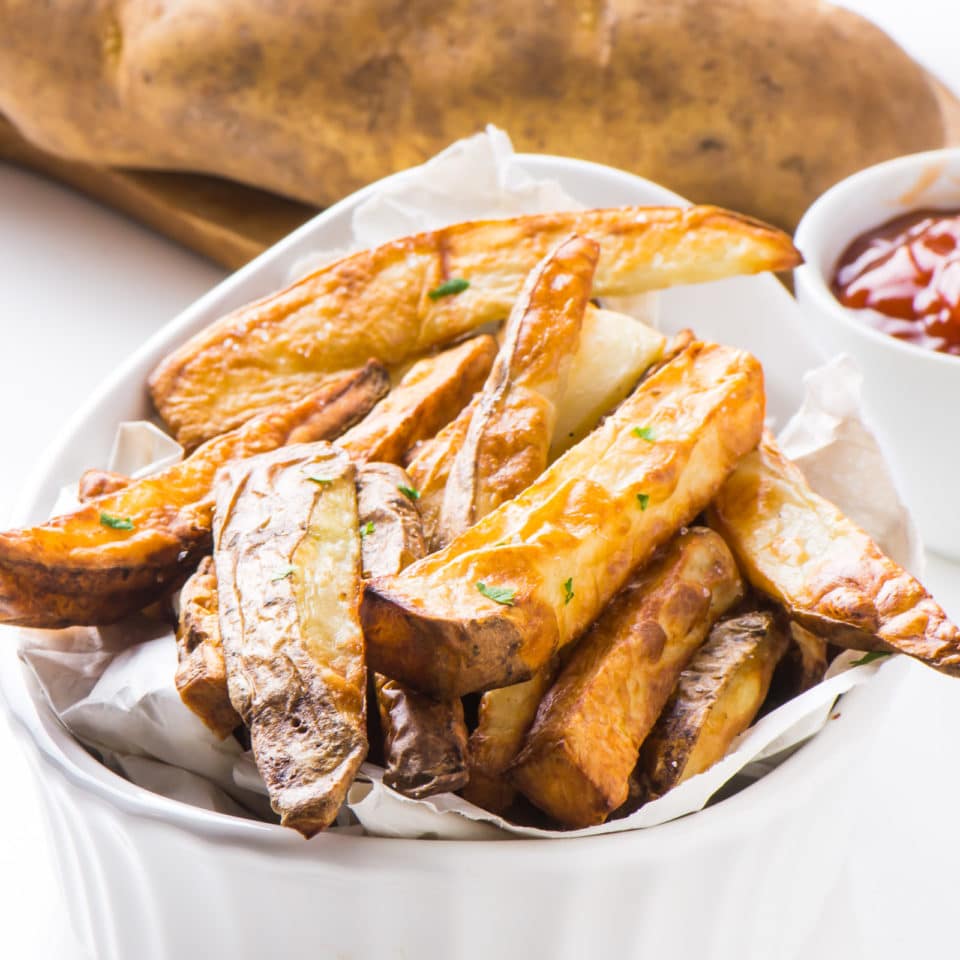 Air Fryer French fries are served up in a white bowl with ketchup and raw potatoes behind it.