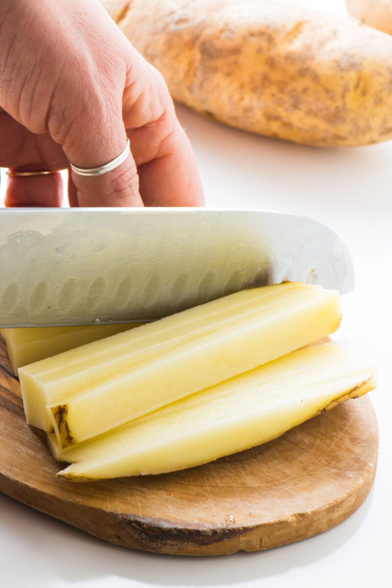 A hand is holding a knife and chopping potatoes in the shape of french fries.