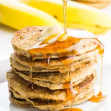 A stack of several vegan banana pancakes sit on a white place with a pat of melty vegan butter on the top. A pitcher is pouring maple syrup over the stack and it is dripping down the sides of the pancakes. There are yellow banana in the background sitting beside another stack of pancakes.
