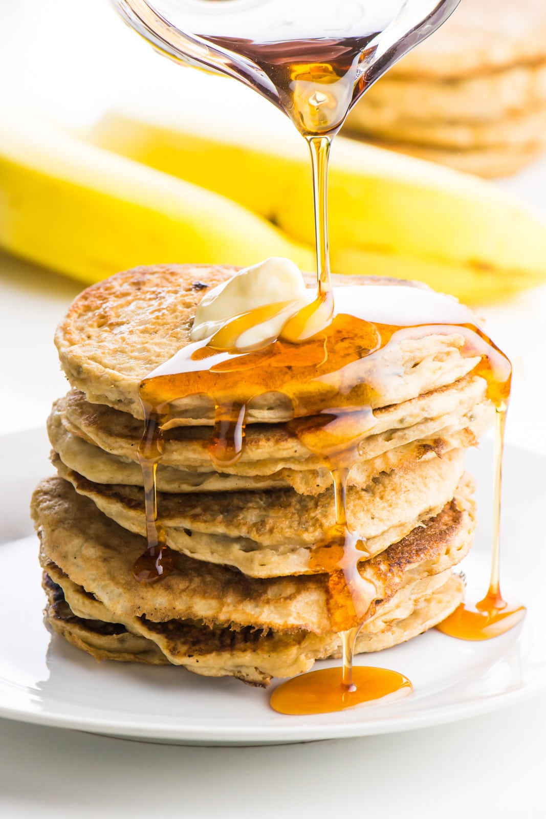 A stack of several vegan banana pancakes sit on a white place with a pat of melty vegan butter on the top. A pitcher is pouring maple syrup over the stack and it is dripping down the sides of the pancakes. There are yellow banana in the background sitting beside another stack of pancakes.