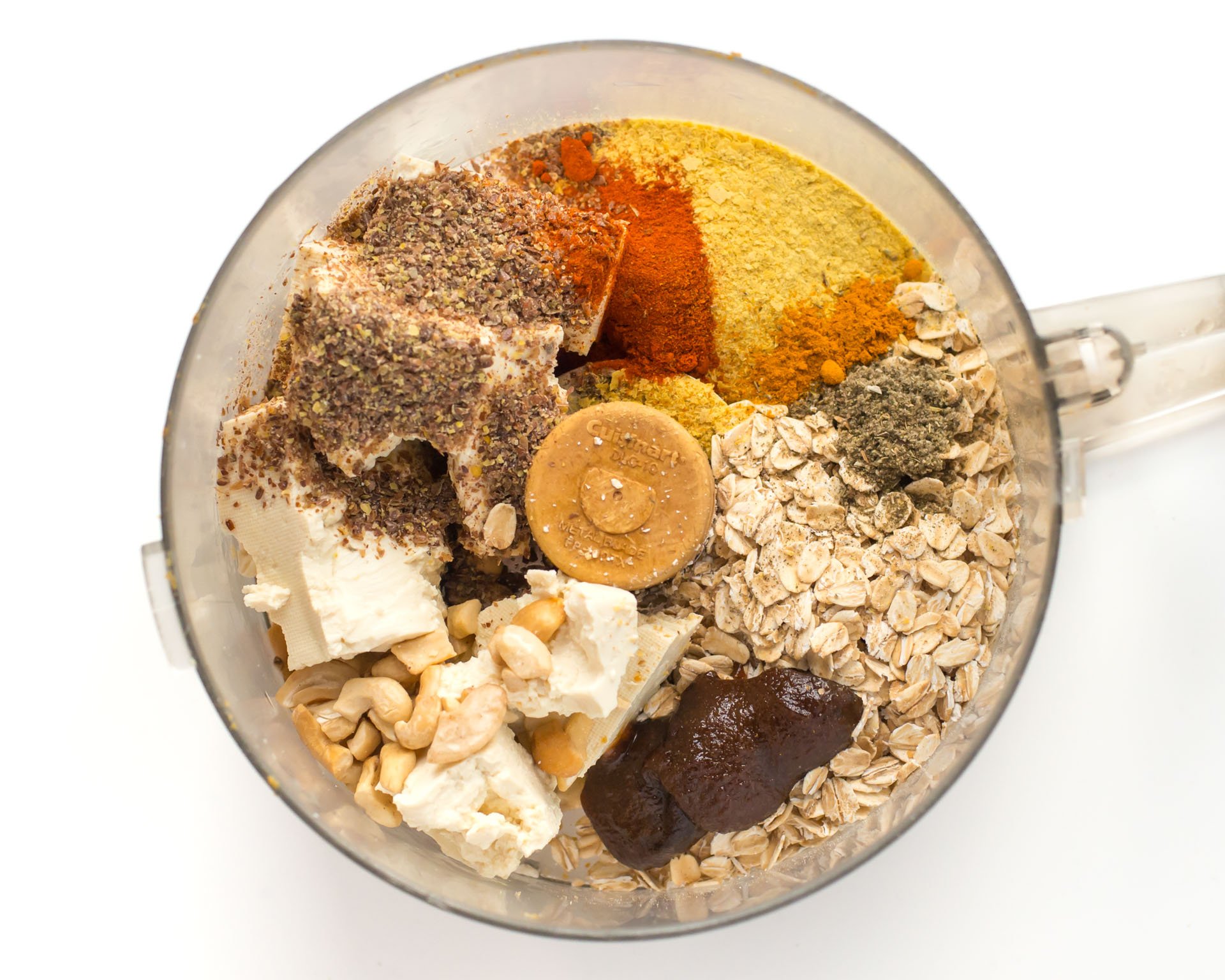 Looking down at a bowl of a food processor with ingredients in it, including oatmeal, tofu, spices, and more.