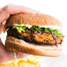 A hand holds a lentil burger, showing off greens and mayo on a whole wheat bun.