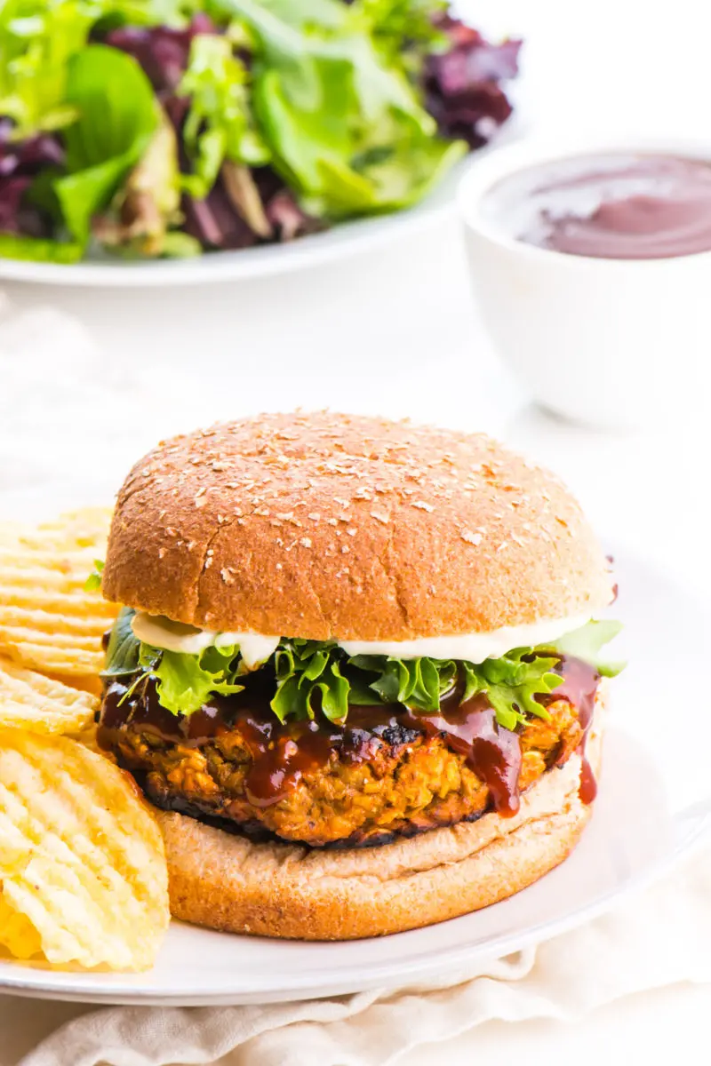 A lentil burger sits on a plate with potato chips beside it. A bowl of sauce and salad greens are behind it.