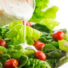 A mason jar full of vegan ranch dressing is pouring the dressing over a leafy, green salad with lots of bright red cherry tomatoes.