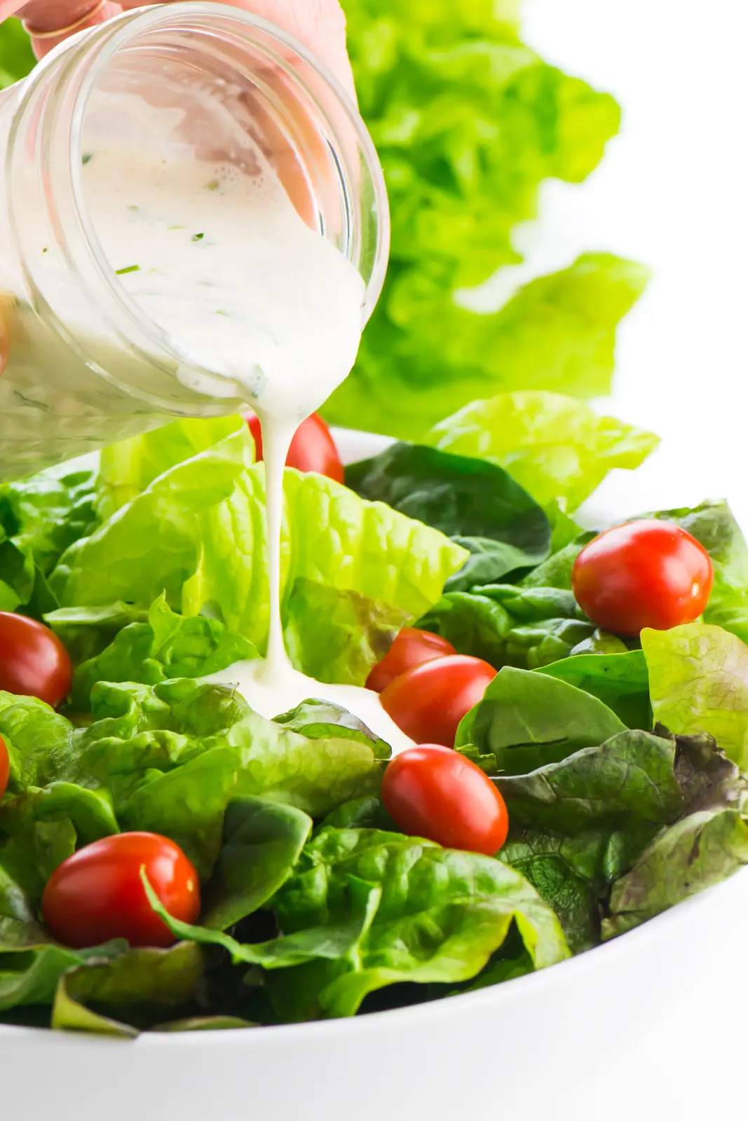 A hand holds a jar of vegan ranch dressing pouring it over a green salad with cherry tomatoes. 