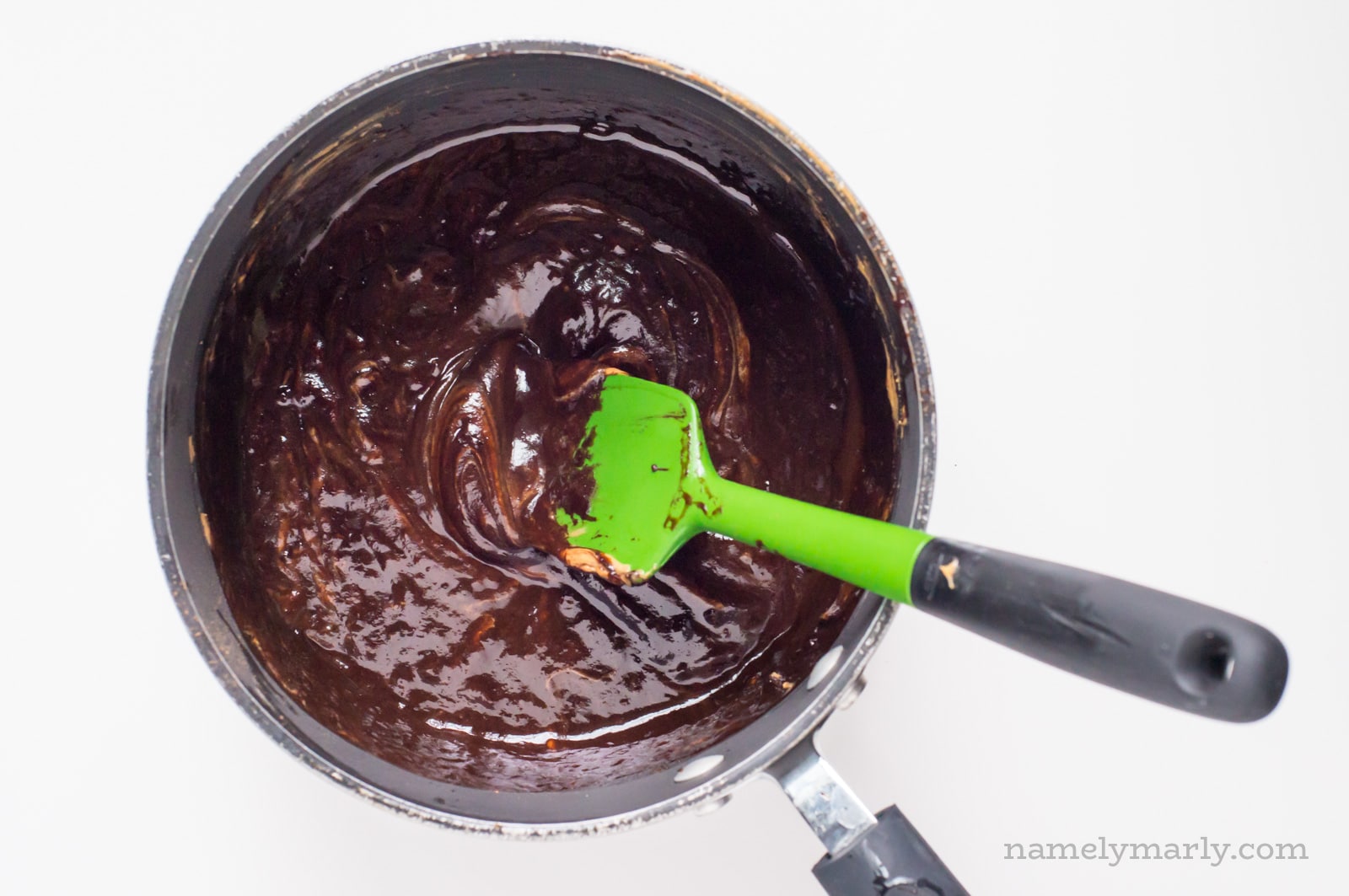 A saucepan is full of chocolate batter. A green spatula is stirring the ingredients.
