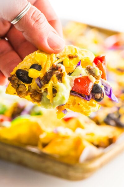 A hand reaches in with a baked vegan nacho taken from the pan behind it.