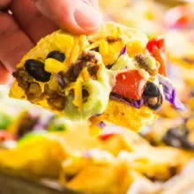 A hand reaches in with a baked vegan nacho taken from the pan behind it.