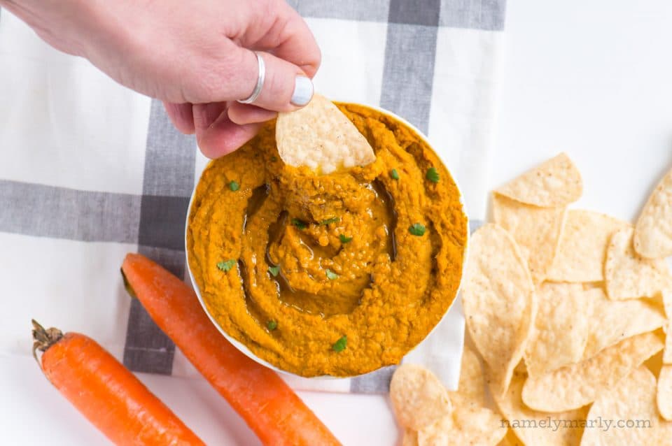 A hand reaches in to dip a chip in a bowl of carrot hummus.