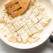 Tofu is in a skillet and it's broken into pieces by a wooden spatula.