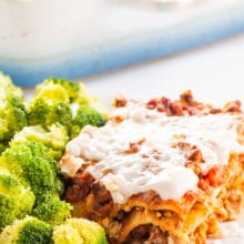 A slice of vegan vegetable lasagna sits next to steamed broccoli. The rest of the lasagna is in a baking dish behind the plate.