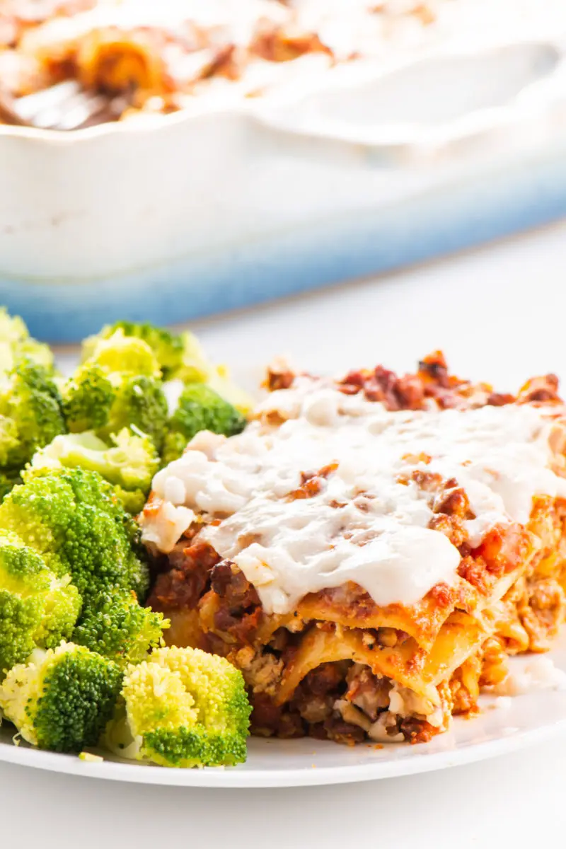 A slice of vegan vegetable lasagna sits next to steamed broccoli. The rest of the lasagna is in a baking dish behind the plate.