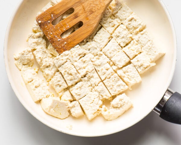 Tofu is in a skillet and it's broken into pieces by a wooden spatula.