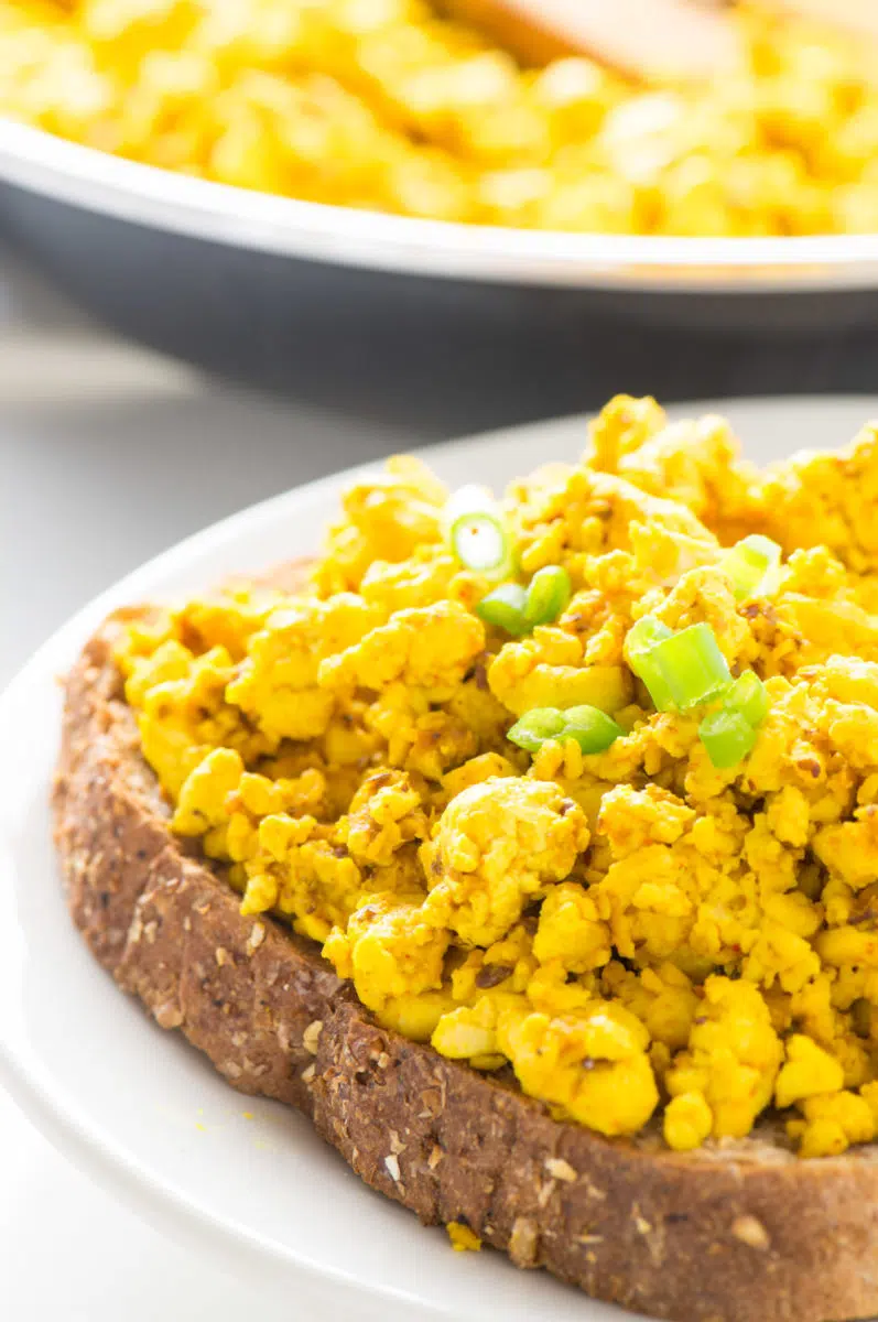 A serving of tofu scramble sits on a piece of toast with sliced green onions. A skillet with more is in the background.
