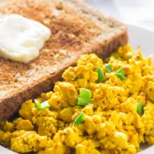 A piece of toast with a pat of melty vegan margarine sits behind a serving of tofu scramble.