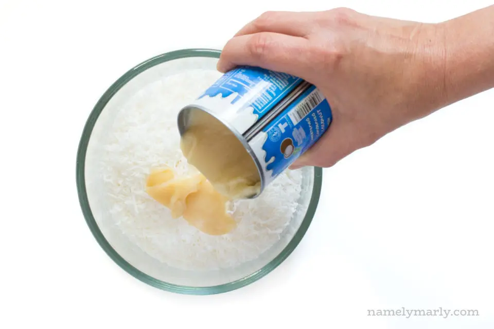 A hand pours coconut sweetened condensed milk into a bowl with shredded coconut.