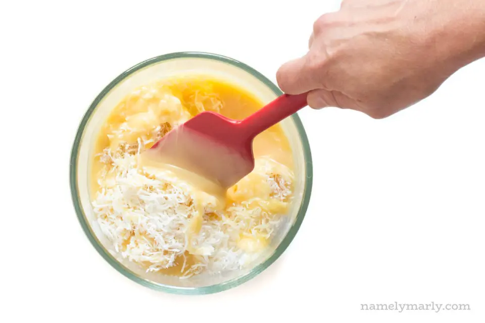 A hand uses a red spatula to stir the sweetened condensed milk and coconut.