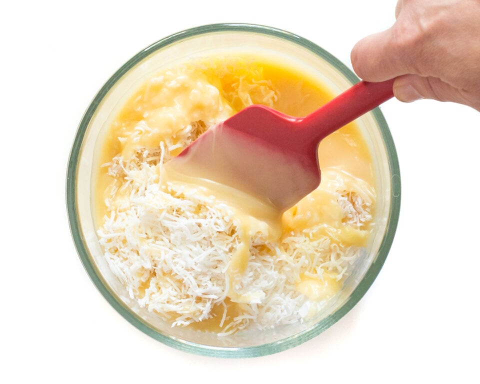 A hand uses a red spatula to stir the sweetened condensed milk and coconut.