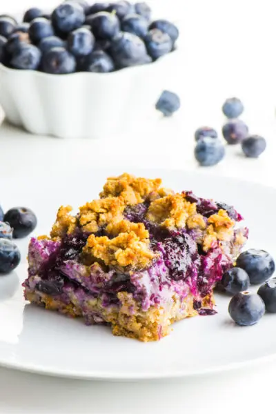 A blueberry oatmeal bar sits on a white plate with lots of blueberries around it.