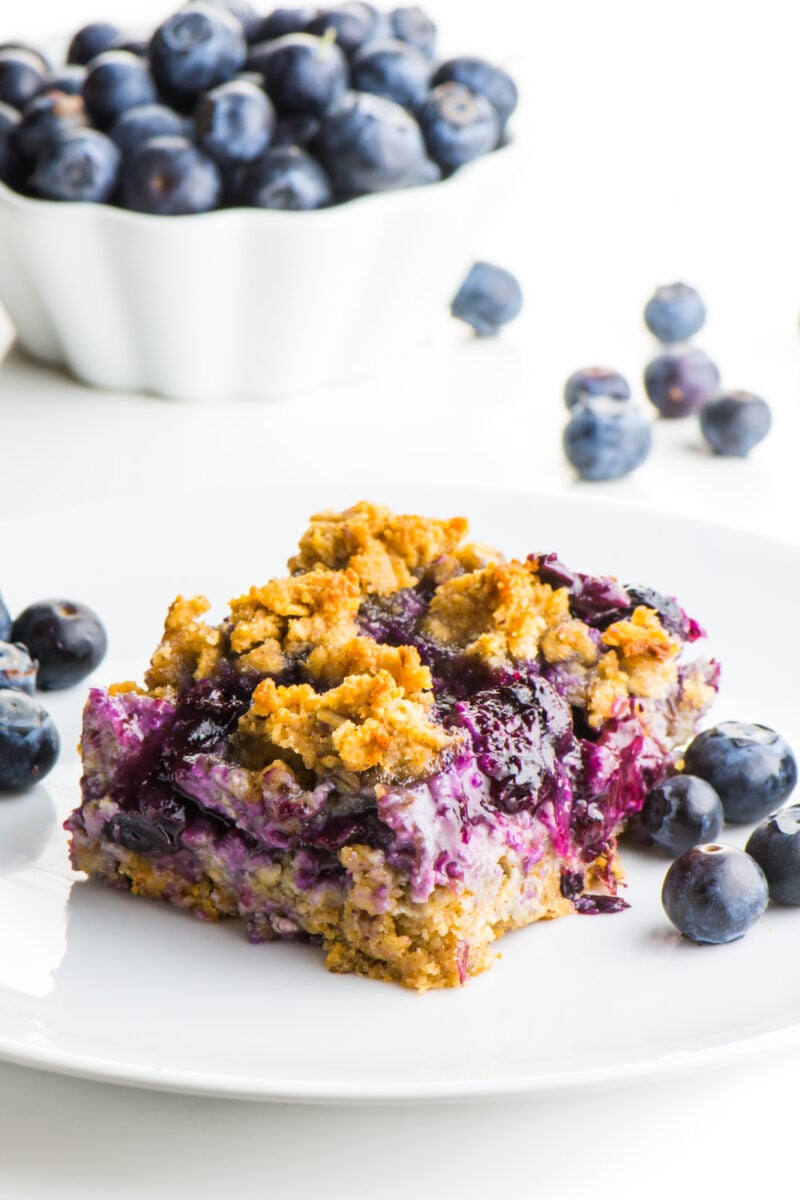 A blueberry oatmeal bar sits on a white plate with lots of blueberries around it.