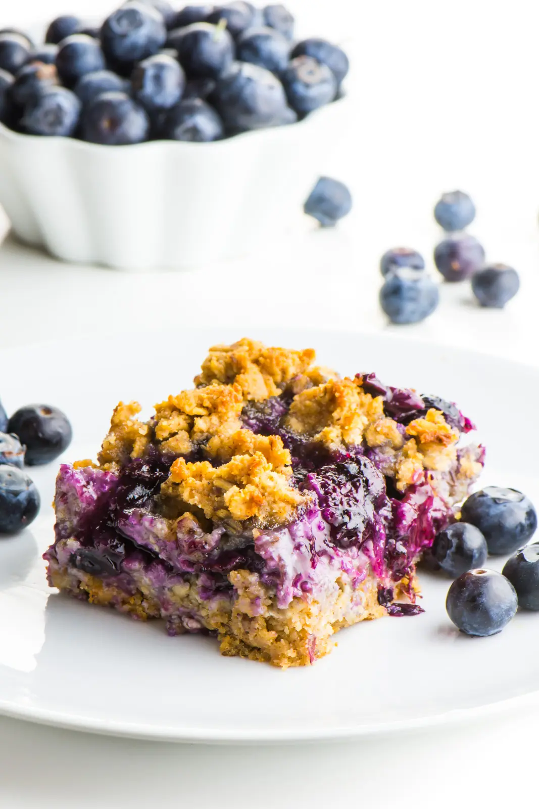 A blueberry crumble bar sits on a plate with lots of blueberries around it.