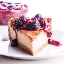A slice of dairy free cheesecake with berry drizzle over the top and more berry topping in a bowl behind it.