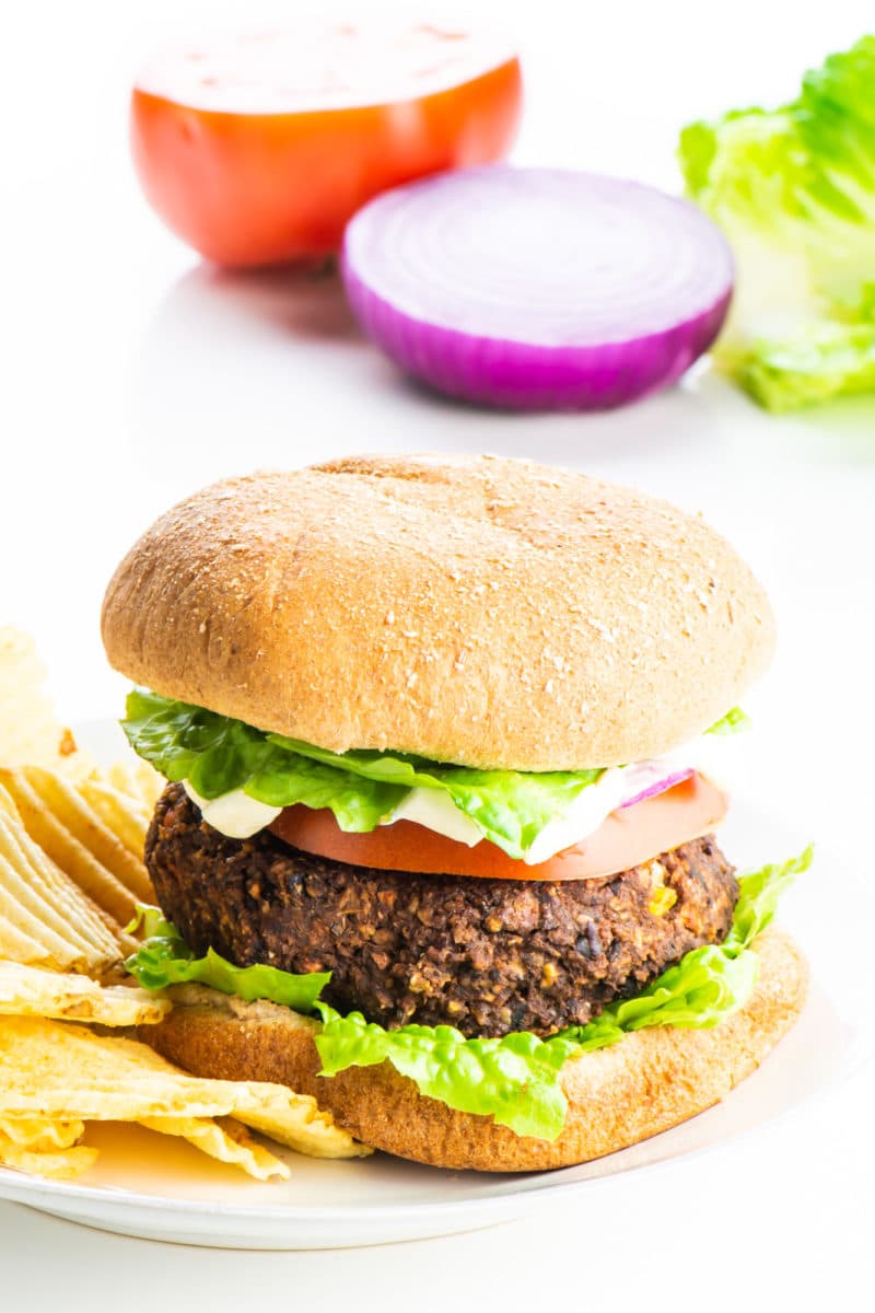 A sweet potato black bean burger sits on a plate with potato chips beside it. Ingredients, like tomatoes and onions, in the background.