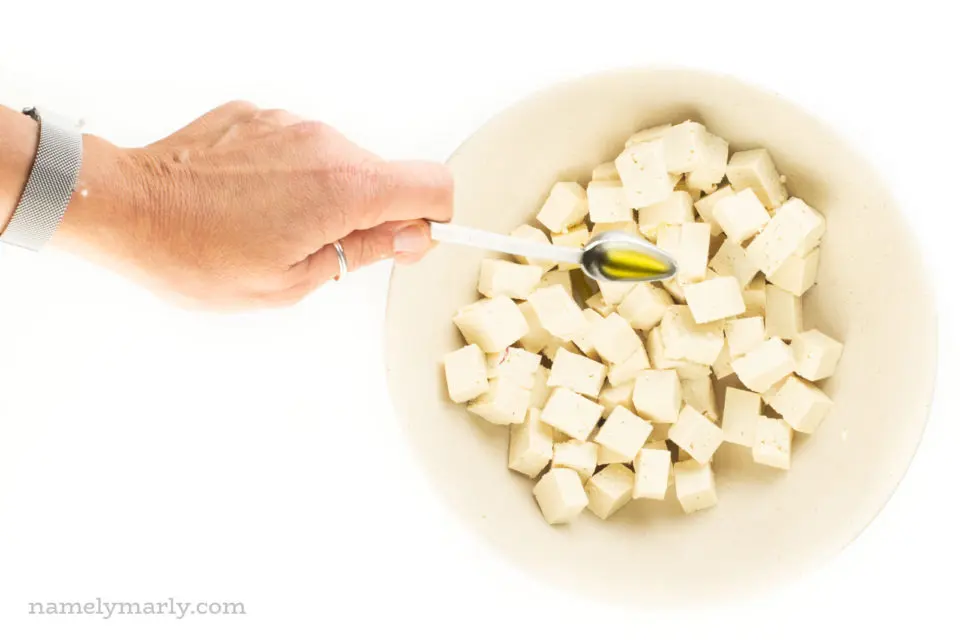 A hand reaches in over a large bowl full of tofu cubes, pouring olive oil.